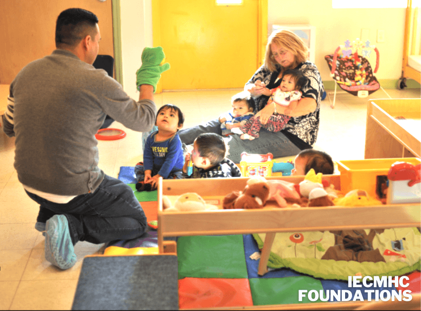 Adults playing with babies and toddlers on the floor of an infant room, captioned IECMHC Foundations.